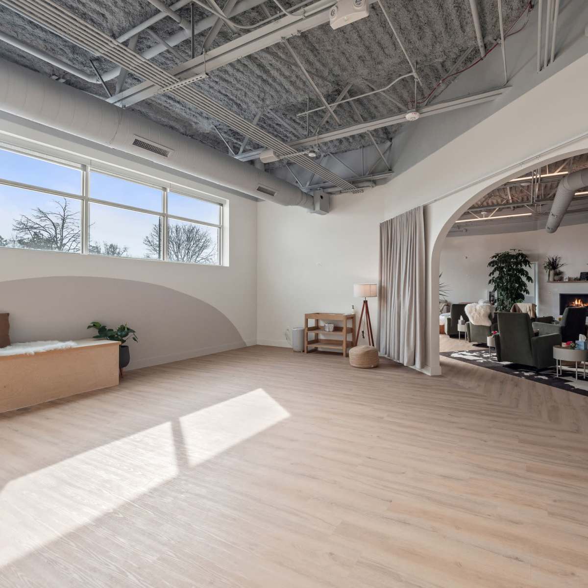 A spacious, well-lit interior with wooden flooring, large windows, and various seating arrangements including a sofa and armchairs.