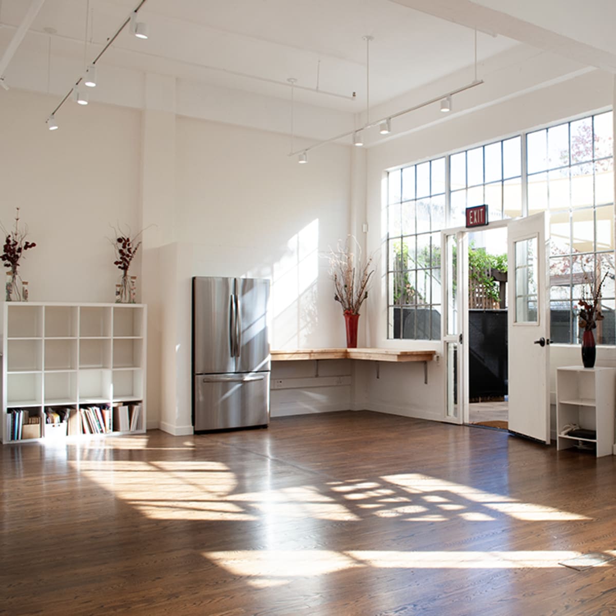 A bright, open interior space with large windows, wooden floors, and minimalist furniture, including a stainless steel refrigerator and a shelf with books.