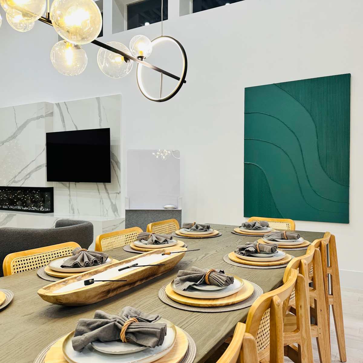 A modern dining room features a long wooden table set for eight, with a large abstract green artwork on the wall and a pendant light above.