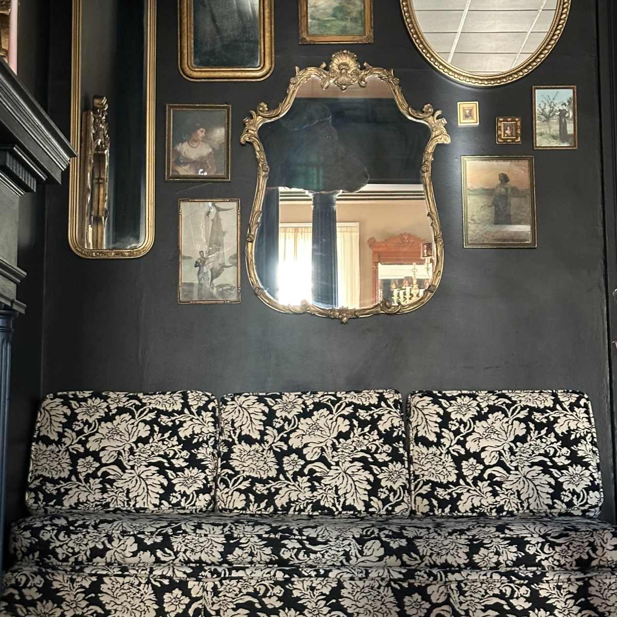A black wall features a variety of ornate mirrors and framed artwork above a floral-patterned sofa.