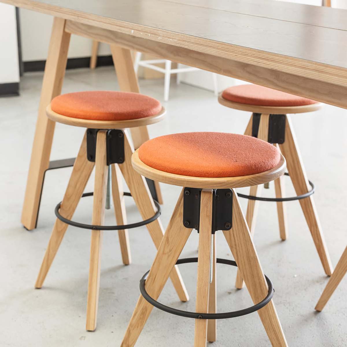 Three wooden stools with orange cushions are arranged around a wooden table in a bright workspace.