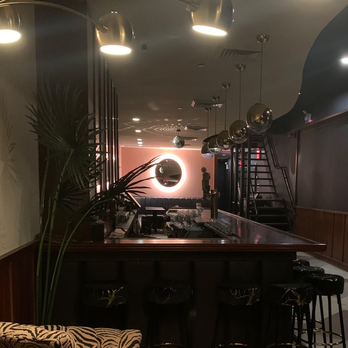 A dimly lit bar area with a counter, several bar stools, and stairs leading to an upper level.