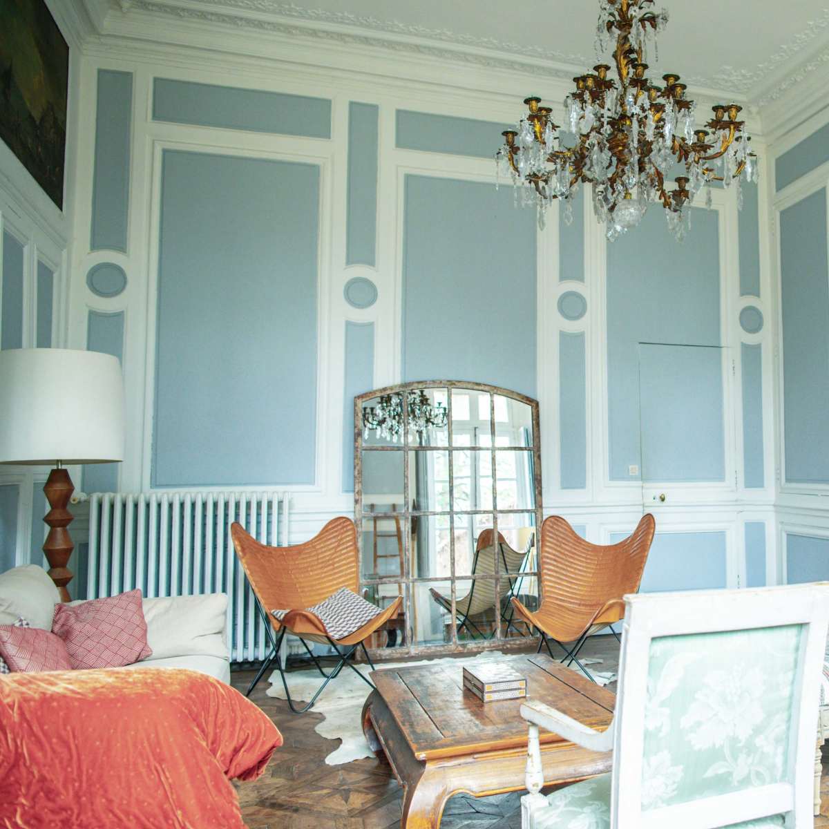 The room features a chandelier, a large mirror, two butterfly chairs, and a marble fireplace surrounded by blue-paneled walls.