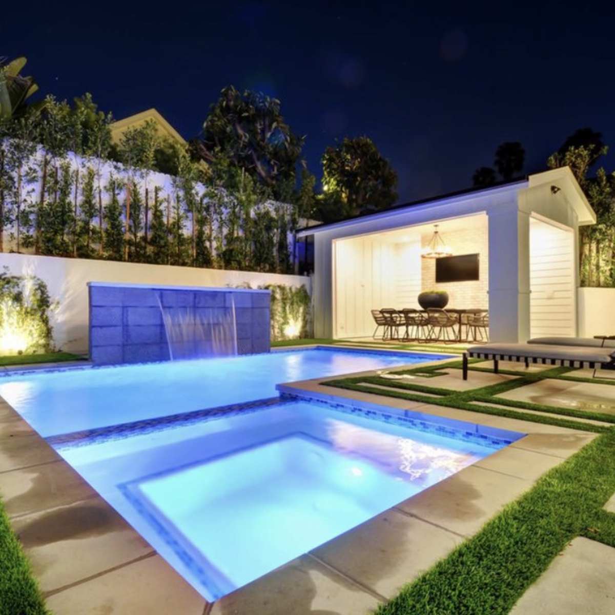 A modern backyard at night featuring a lit swimming pool with a waterfall, surrounded by green landscaping and a small seating area.