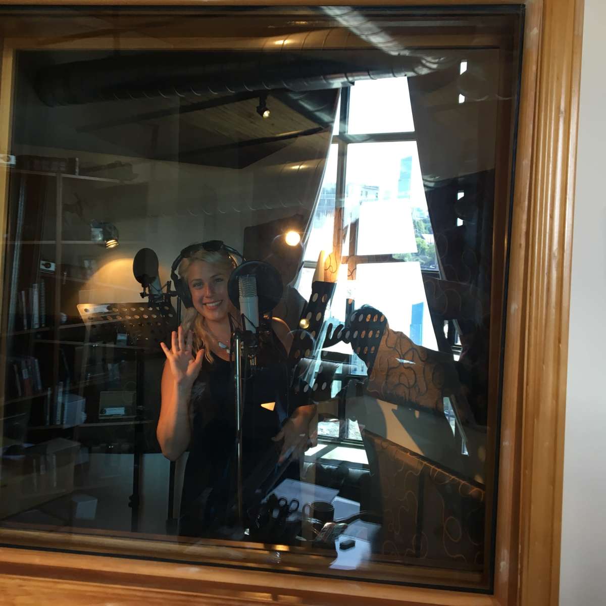 A person stands inside a sound recording studio, smiling and gesturing while microphone equipment surrounds them.