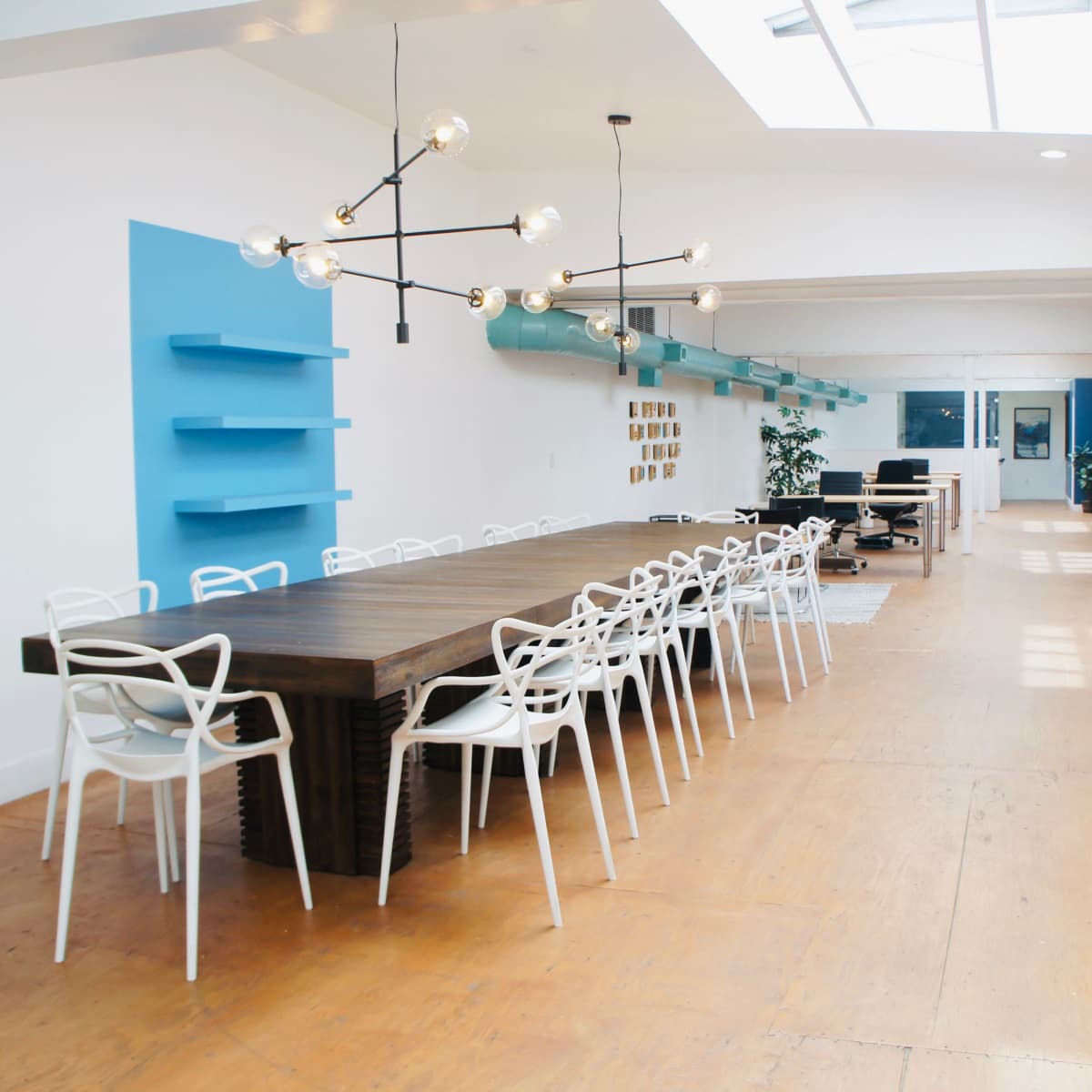 A long wooden table with white chairs is centered in a bright, open room featuring a blue accent wall and large windows.