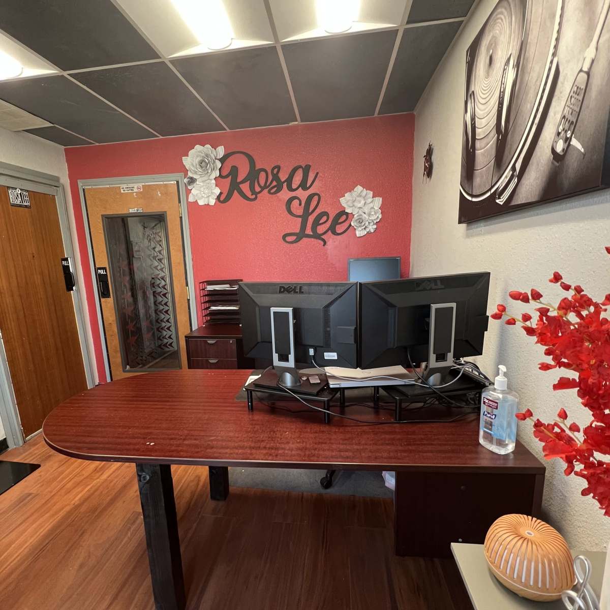 An office reception area featuring a wooden desk with two Dell monitors, a hand sanitizer dispenser, and decorative elements on the walls.
