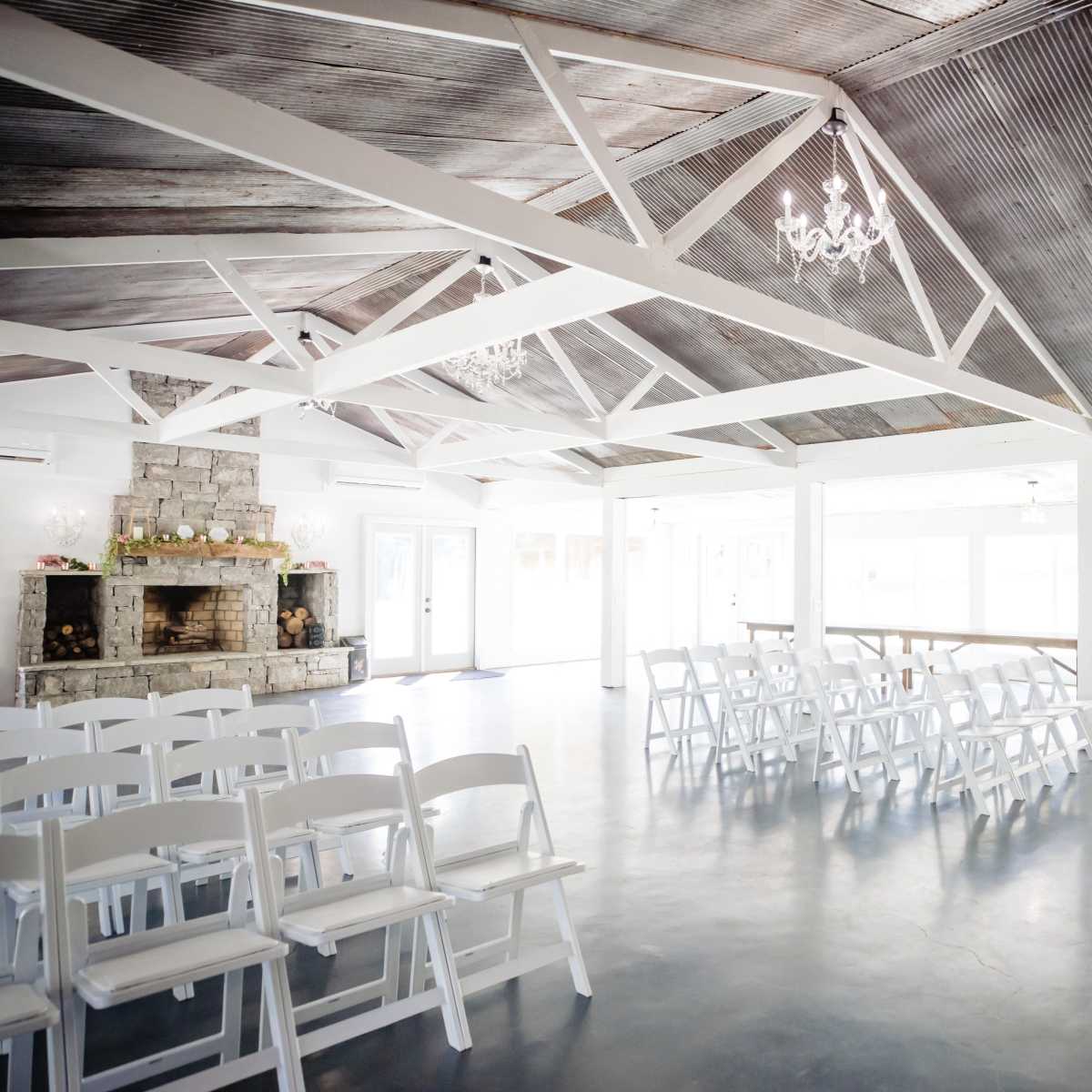 The interior of a spacious event venue features white folding chairs arranged facing a central area, with a stone fireplace and chandeliers enhancing the rustic ceiling.