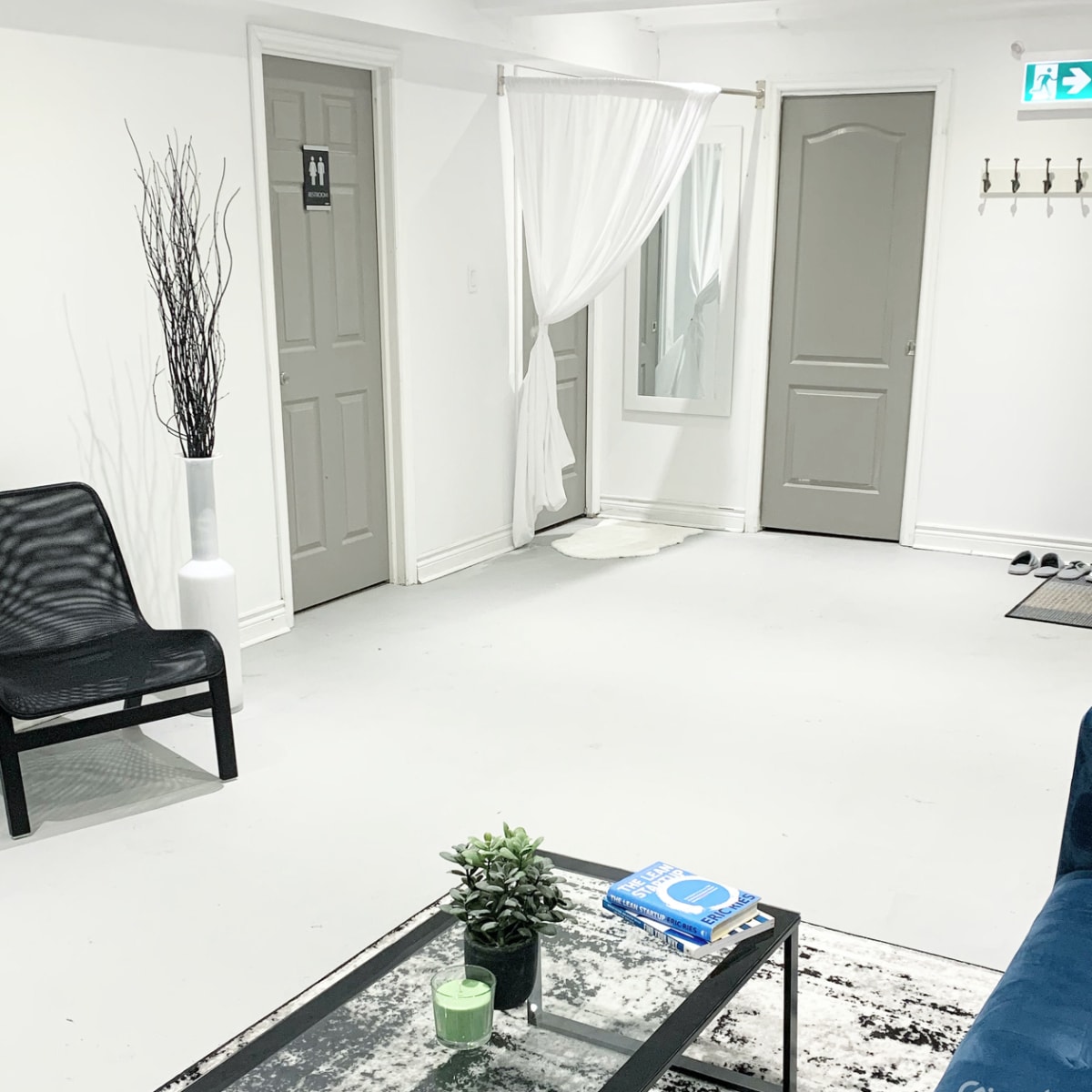 A minimalist waiting area with a chair, a small coffee table, a potted plant, and a mirror, along with two doors and a curtain in a bright, neutral-colored room.