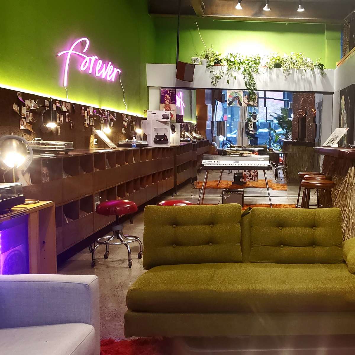 The scene features a retro-styled lounge area with a green sofa in the foreground, surrounded by a vibrant, eclectic space that includes a bar, musical instruments, and decorative plants.