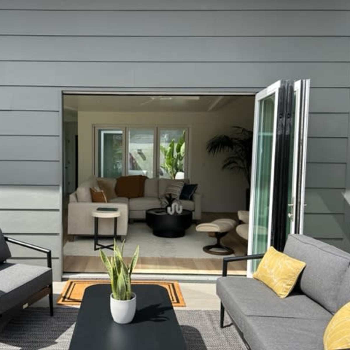 A modern outdoor seating area is viewed through an open doorway, leading into a light-filled living space with contemporary furniture.