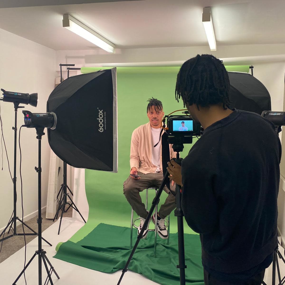 A person is being filmed in front of a green backdrop with studio lighting in a photography setup.
