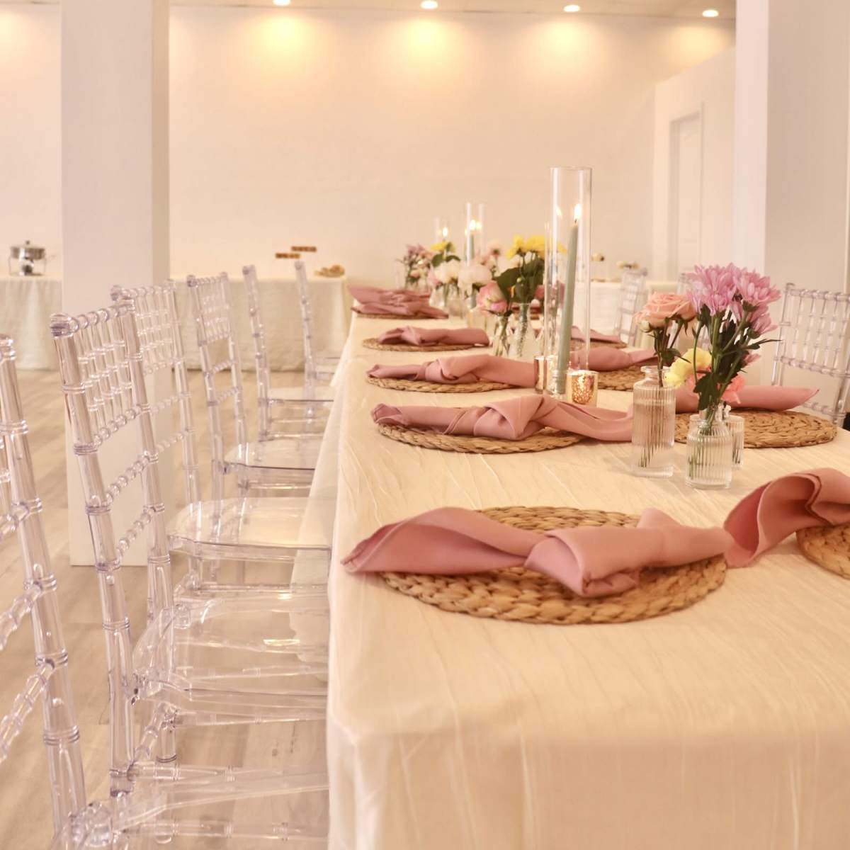 A long dining table is set with pink napkins, woven placemats, and clear chairs, adorned with floral centerpieces and candles.