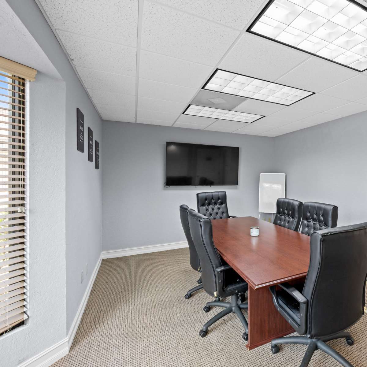 A conference room with a rectangular wooden table surrounded by six black leather chairs, a television mounted on the wall, and a whiteboard in the corner.