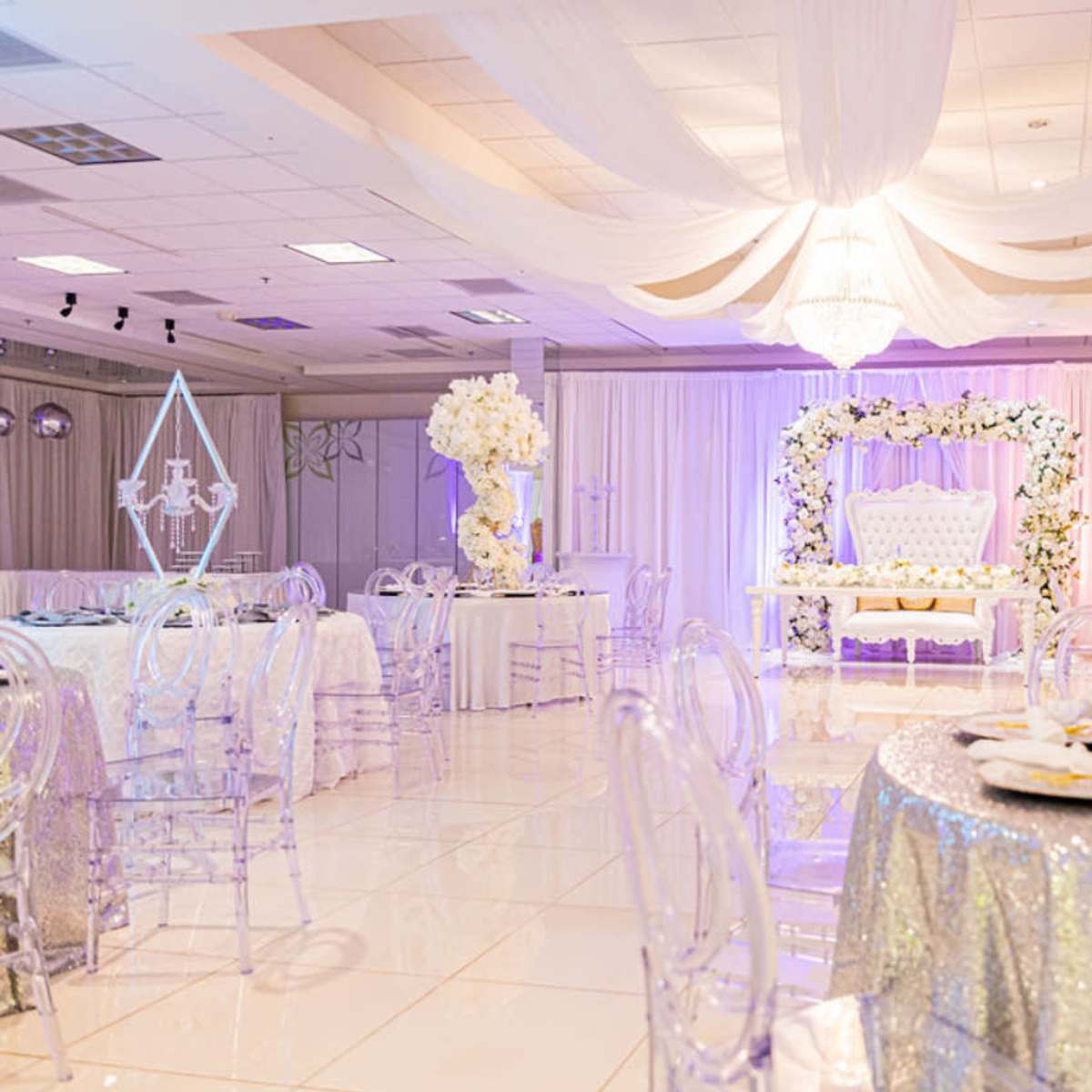 The scene shows a banquet hall decorated with elegant tables, transparent chairs, and floral arrangements, featuring a large backdrop with a throne and draping overhead.