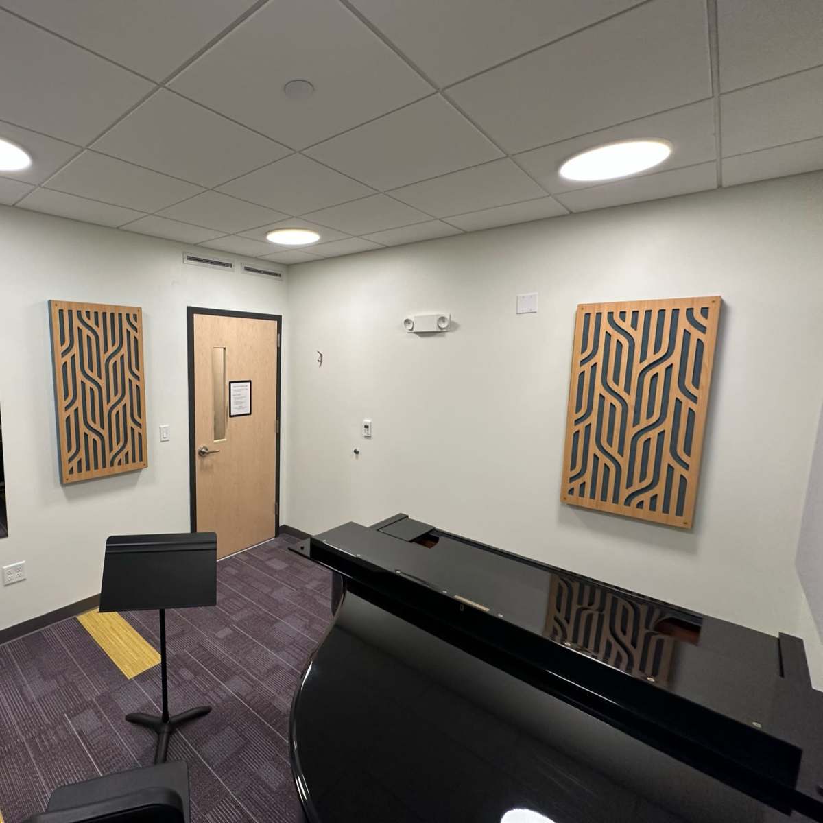 A small, well-lit music practice room featuring a grand piano, sound-absorbing wall panels, and a music stand.