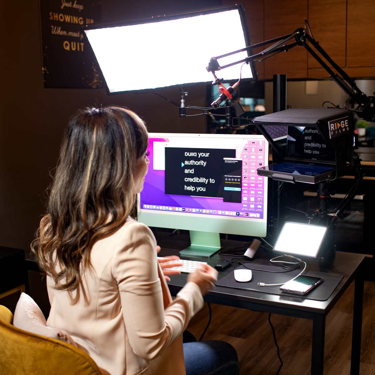 A person is sitting at a desk using a computer, with overhead lighting and filming equipment set up in a modern indoor space.