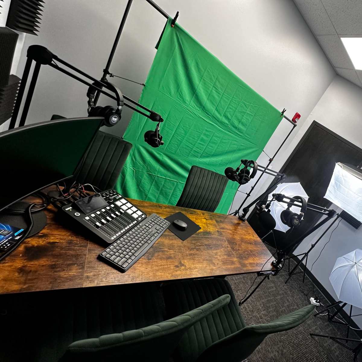 A recording studio setup features a wooden table with a keyboard and mouse, green screen backdrop, and two softbox lights.
