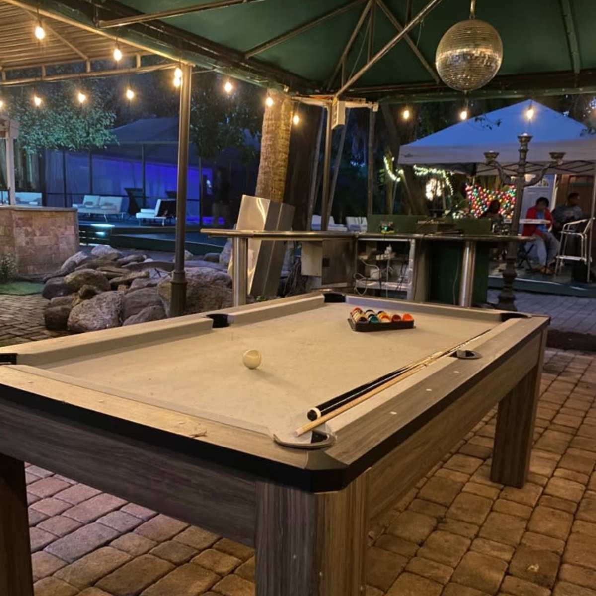A pool table with an unfinished game sits under a canopy adorned with string lights in an outdoor recreational area.