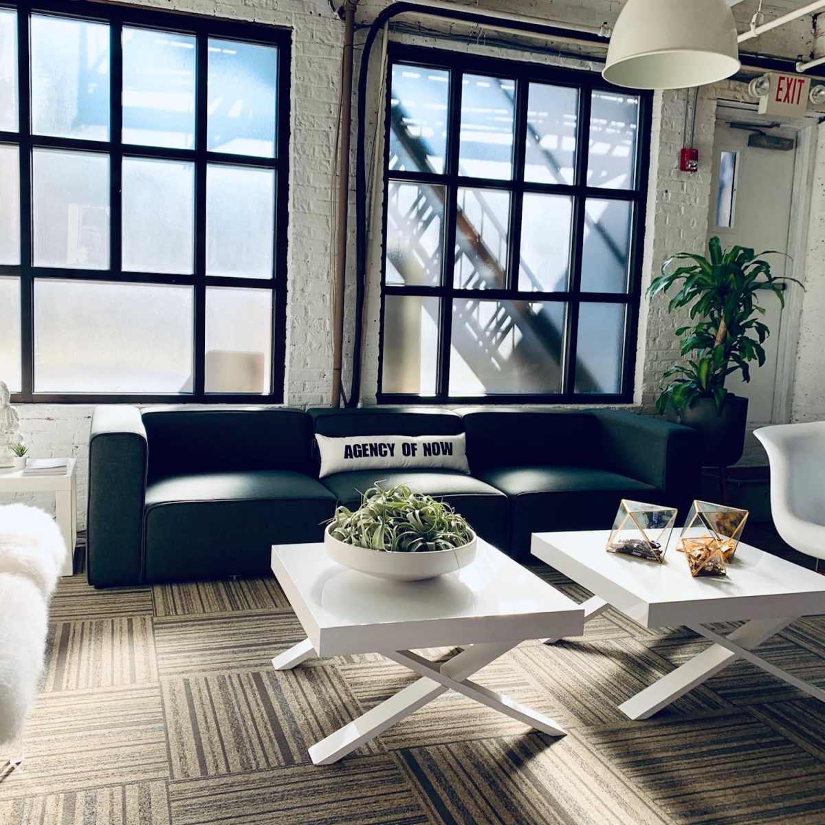 A modern office lounge area featuring a dark green sofa, white coffee tables, and large windows, with plants and decorative items.