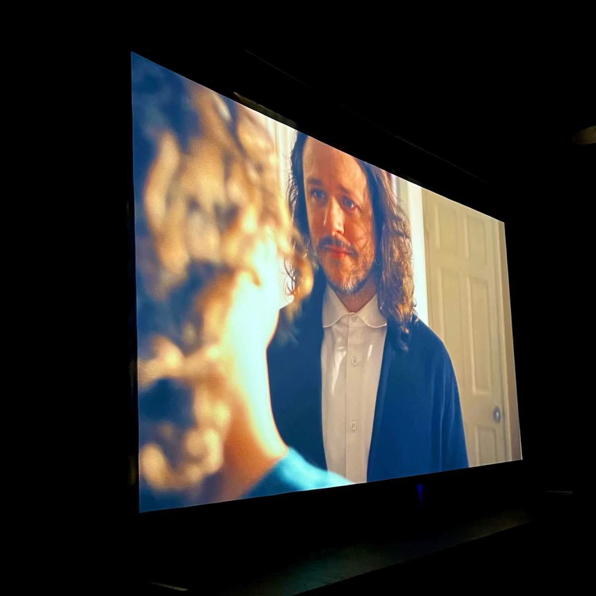 A projection of a man with long hair and a beard speaks to a child in a dimly lit room.