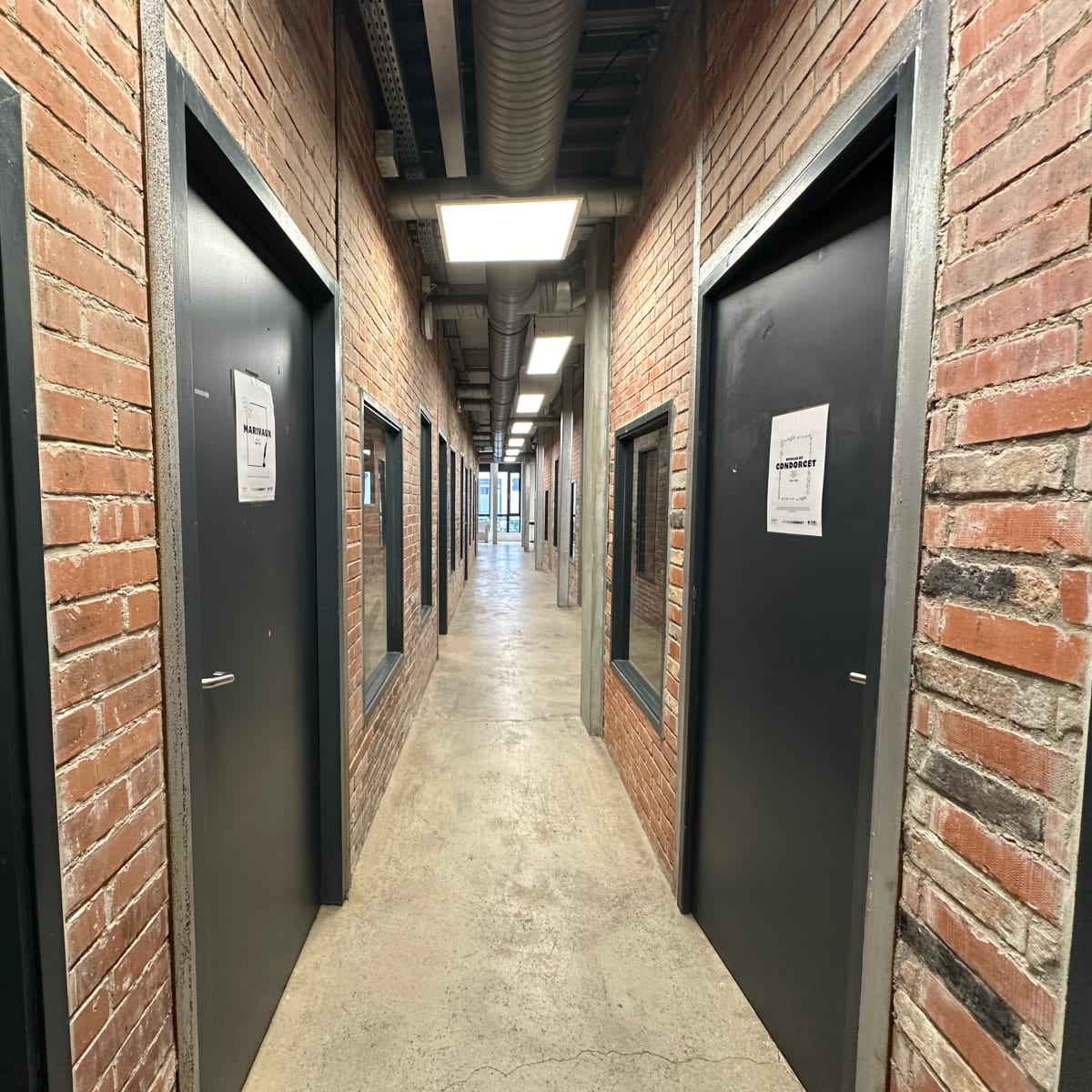A narrow corridor lined with dark doors and exposed brick walls, leading to a well-lit area at the end.