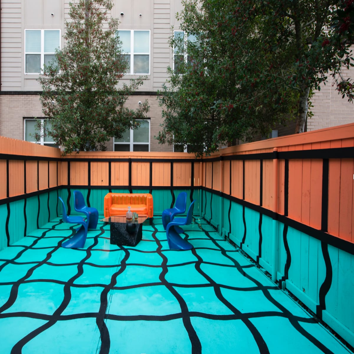 A brightly painted outdoor space with a turquoise floor featuring a black grid pattern, orange benches, and blue chairs surrounded by wooden fencing.