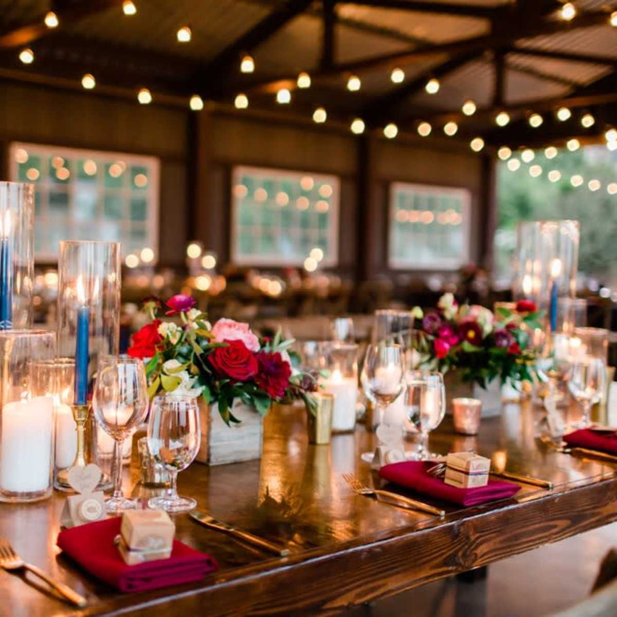 A beautifully arranged dining table is set with flowers, candles, and glassware under string lights in a rustic indoor venue.