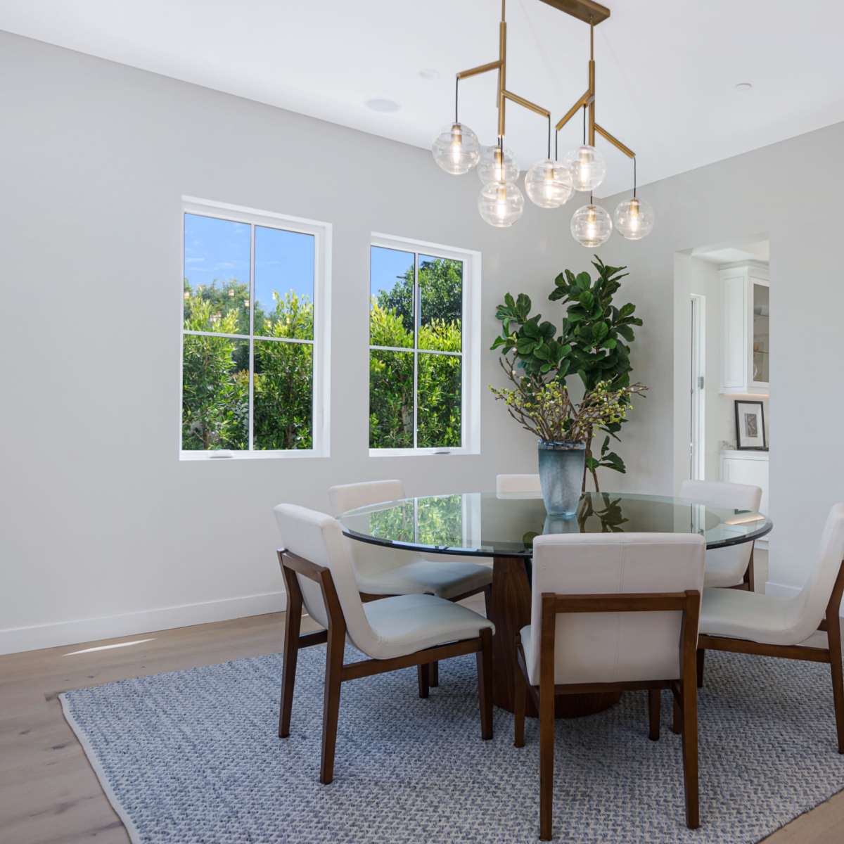 A circular glass dining table with eight chairs is set in a modern room featuring large windows and a decorative wall piece.