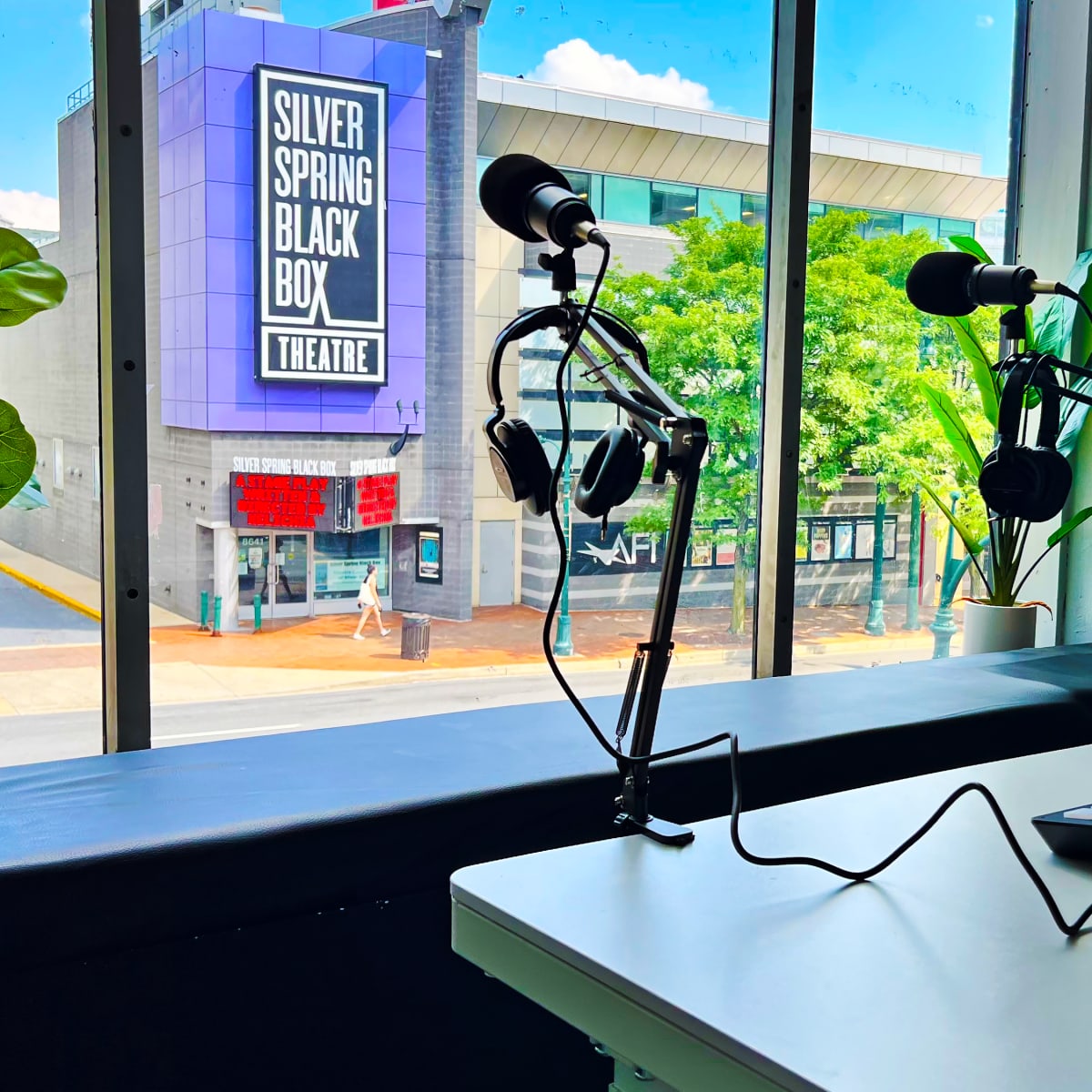 A recording studio setup with a microphone and headphones on a table, overlooking a street with the Silver Spring Black Box Theatre visible through the window.