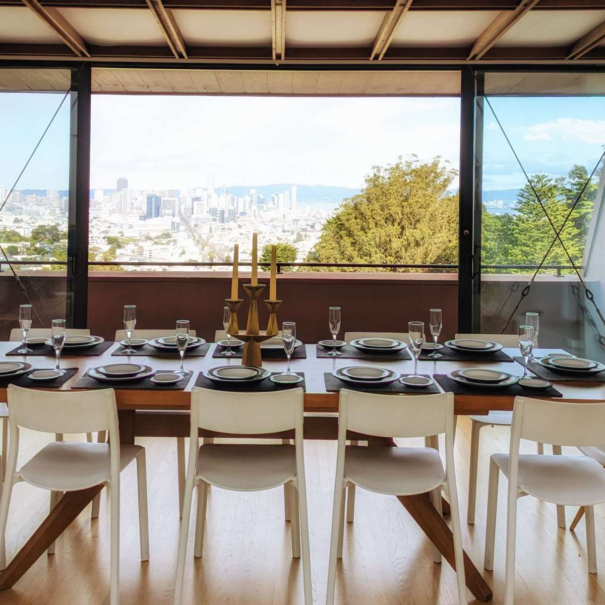 A long wooden dining table is set with plates and glasses, facing large windows that overlook a city skyline.