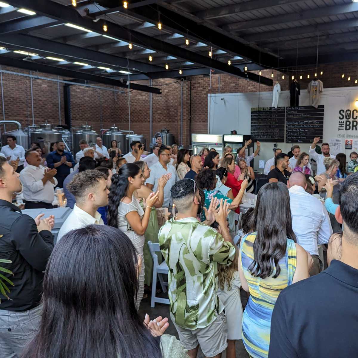 A large group of people is gathered in a brewery, clapping and facing a speaker at the front of the room.