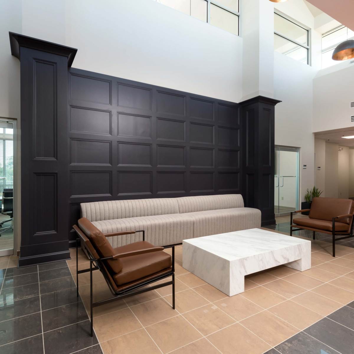 A modern lobby area featuring a gray sofa, two brown arm chairs, and a marble coffee table, all set against a dark paneled wall.
