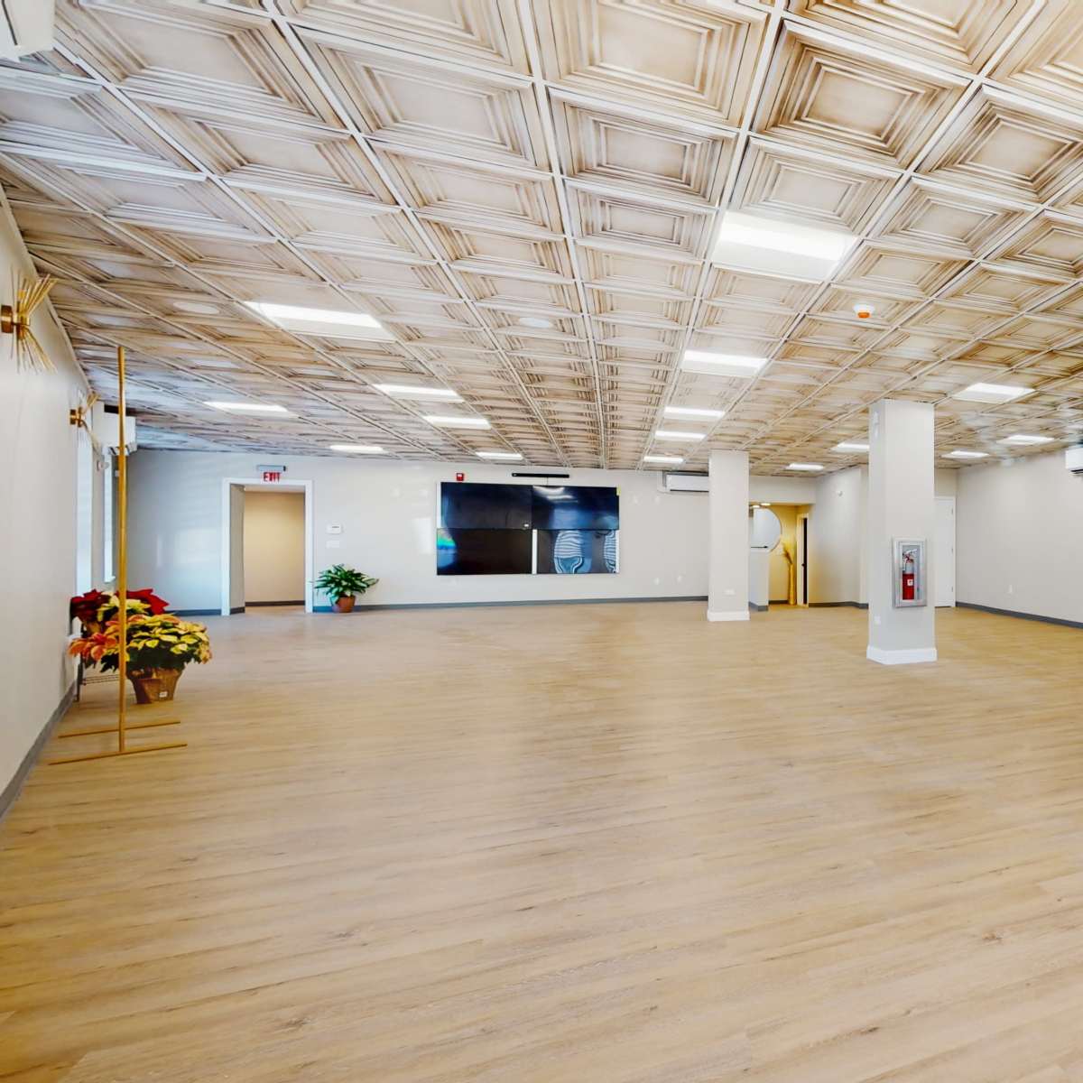 A large, empty room with wooden flooring, high ceilings featuring decorative tiles, and several support pillars.