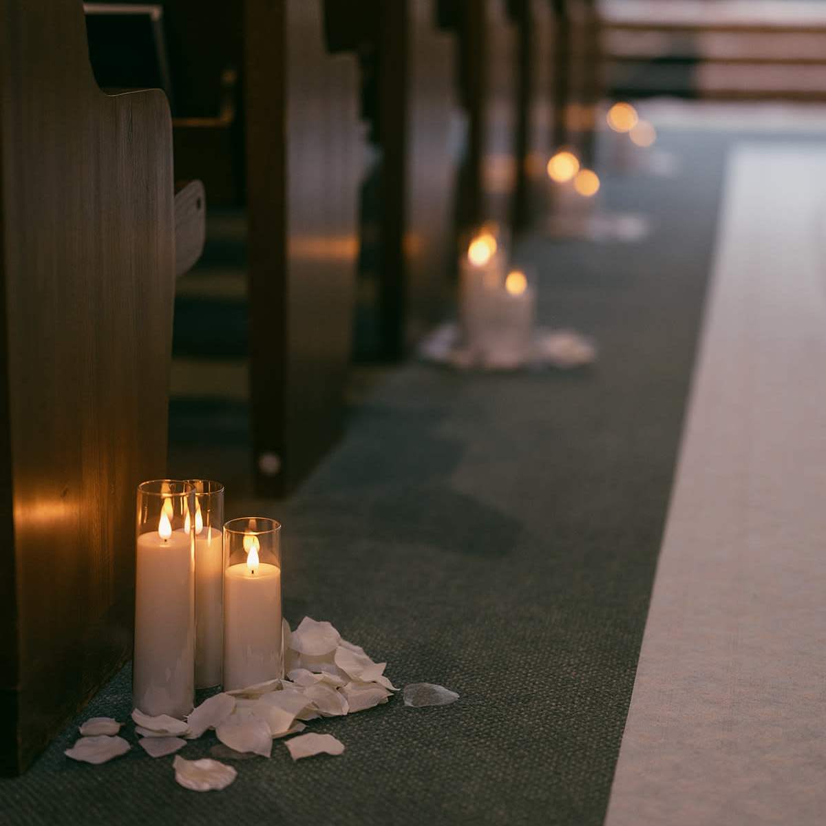 A row of church pews with lit candles and rose petals arranged along the aisle.