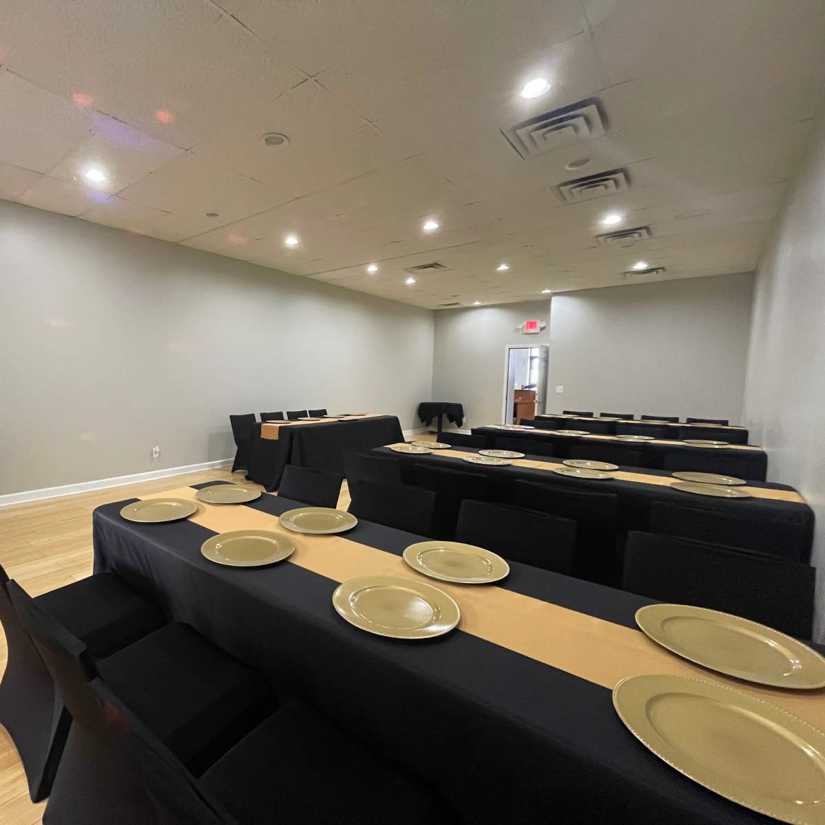 A banquet hall is set up with long tables covered in black cloth and adorned with gold plates, ready for an event.