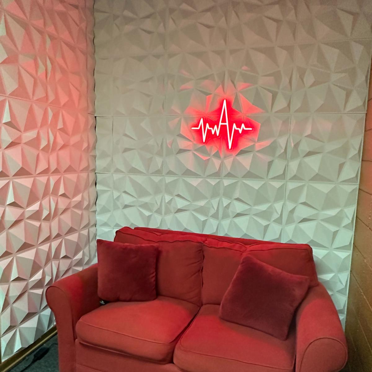 A red couch with two cushions in a corner of a room featuring textured white walls and a red heart rate neon sign above it.