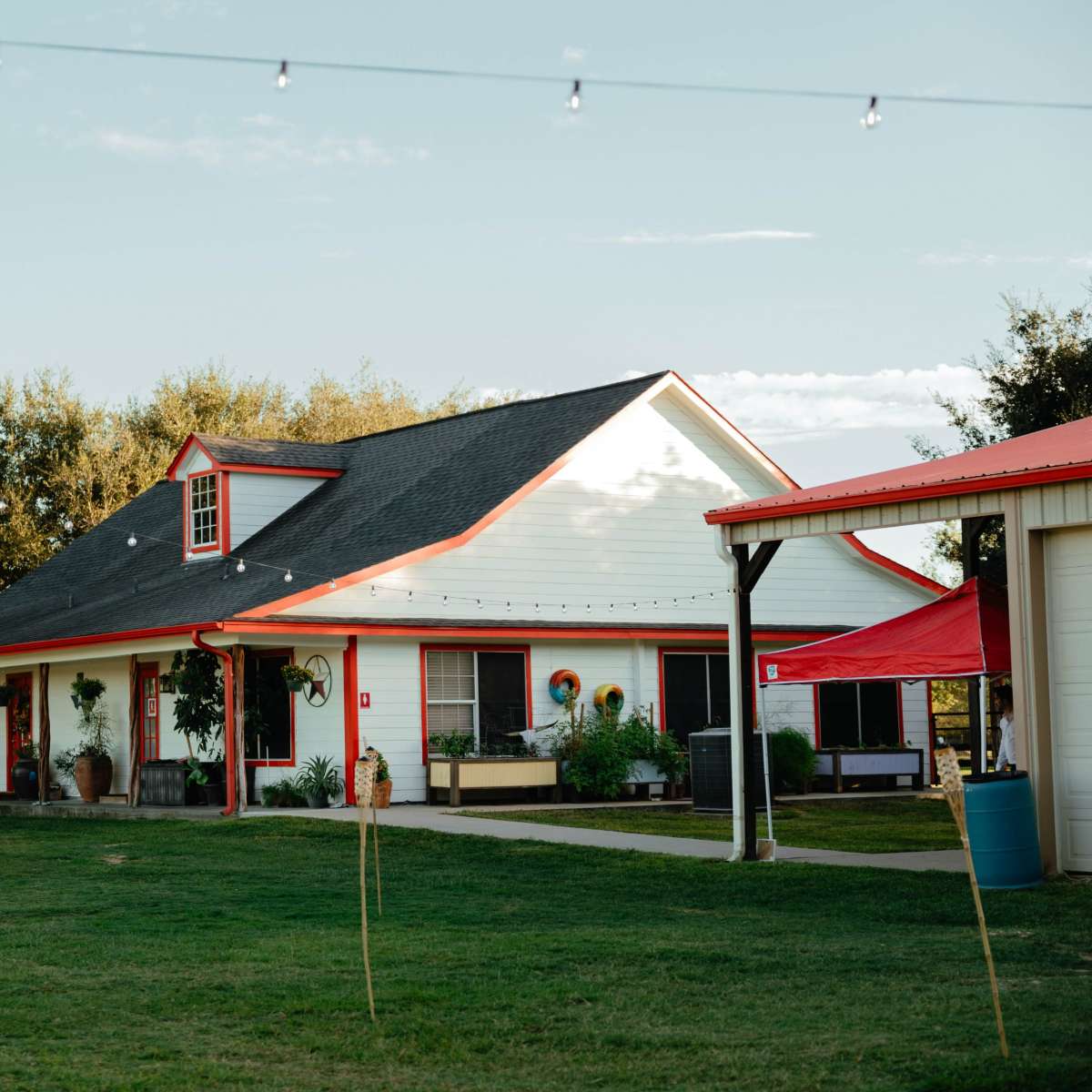 A white house with red accents sits in a grassy area, surrounded by trees and string lights.
