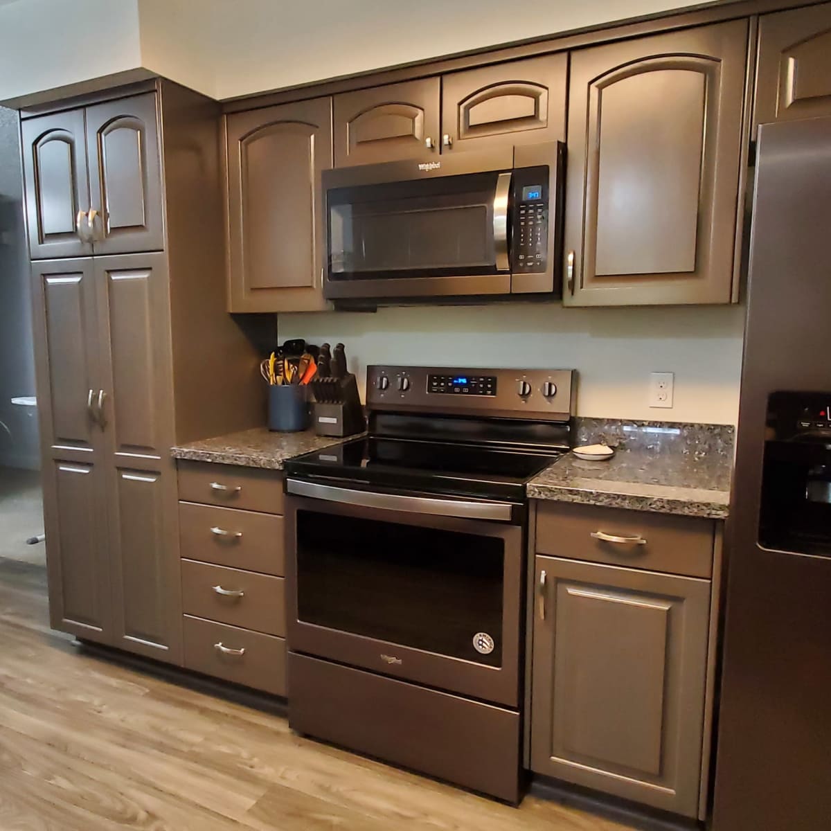 A modern kitchen with dark cabinets, a stainless steel refrigerator, a microwave over the stove, and a dining area in the background featuring a wooden table and chairs.