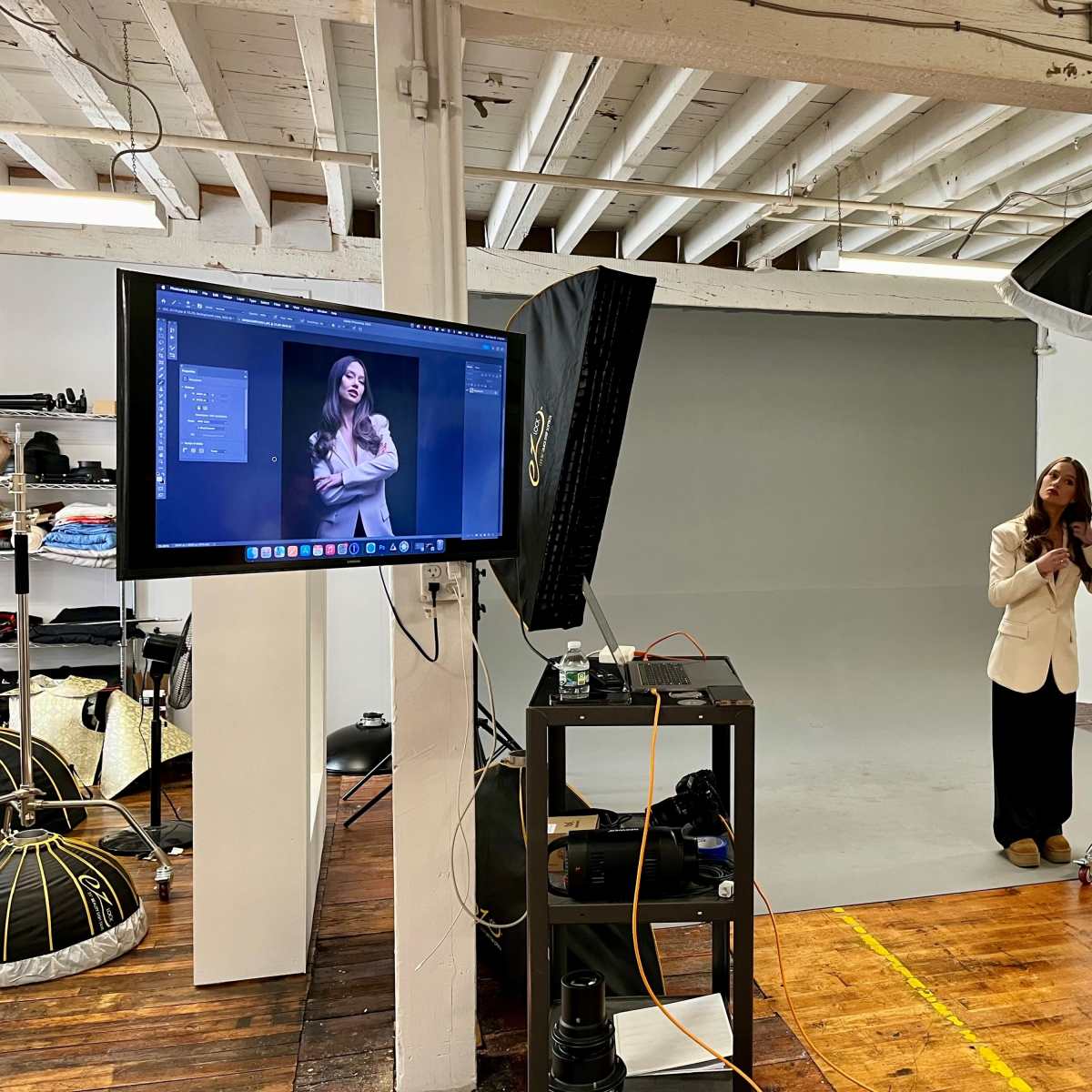 A woman in a white blazer poses in a photography studio with a gray backdrop while a monitor displays her image.