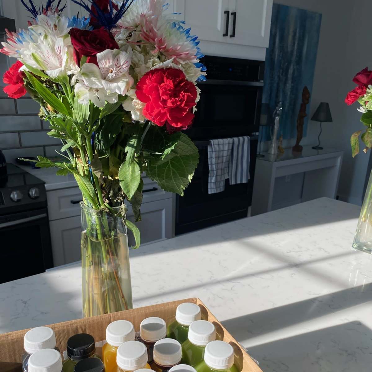 A bouquet of flowers sits in a vase on a kitchen counter next to a box of colorful bottled drinks.