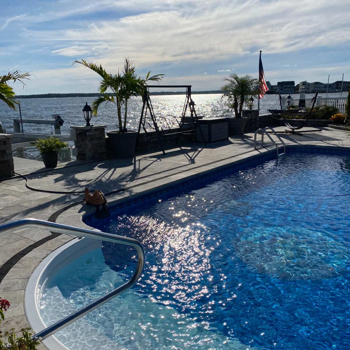 A swimming pool with clear blue water, surrounded by palm trees and a view of the water in the background under a partly cloudy sky.