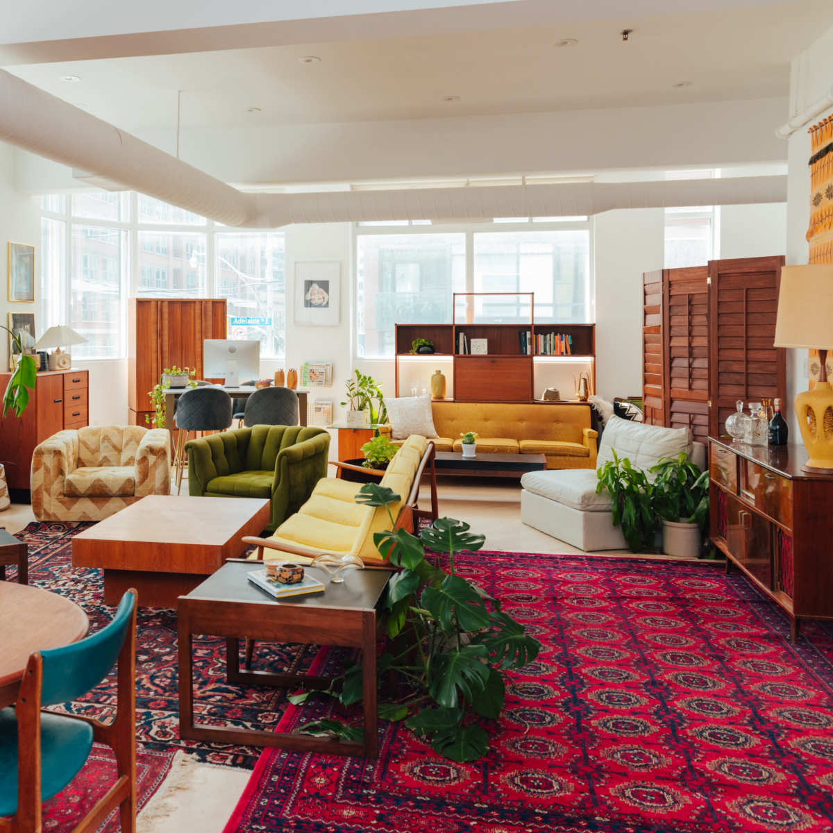 A spacious, well-lit living area featuring various furniture pieces, including a large dining table, several couches, and potted plants on a patterned rug.