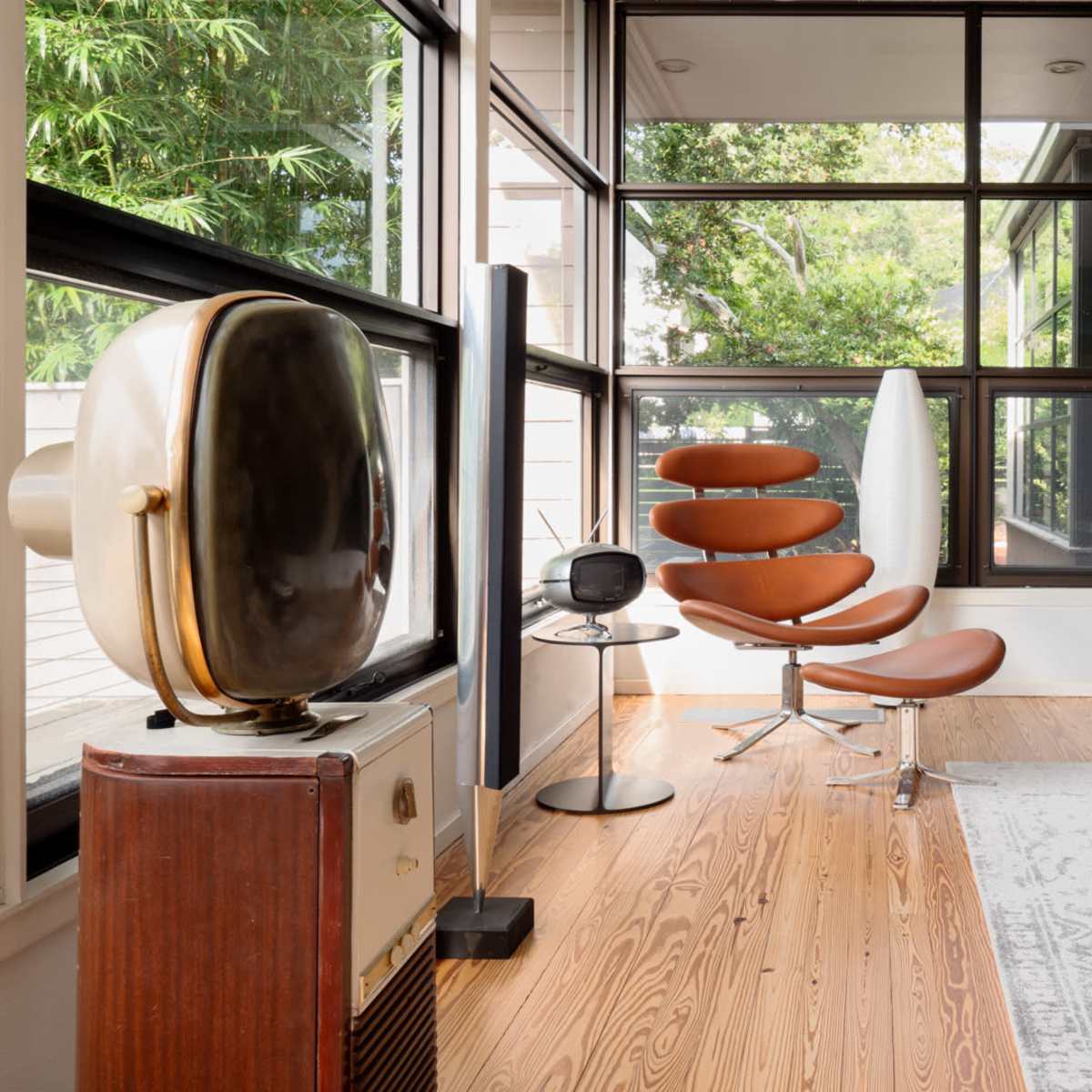 A modern interior with large windows, featuring a brown lounge chair, a sleek standing lamp, and a vintage television set on a table.