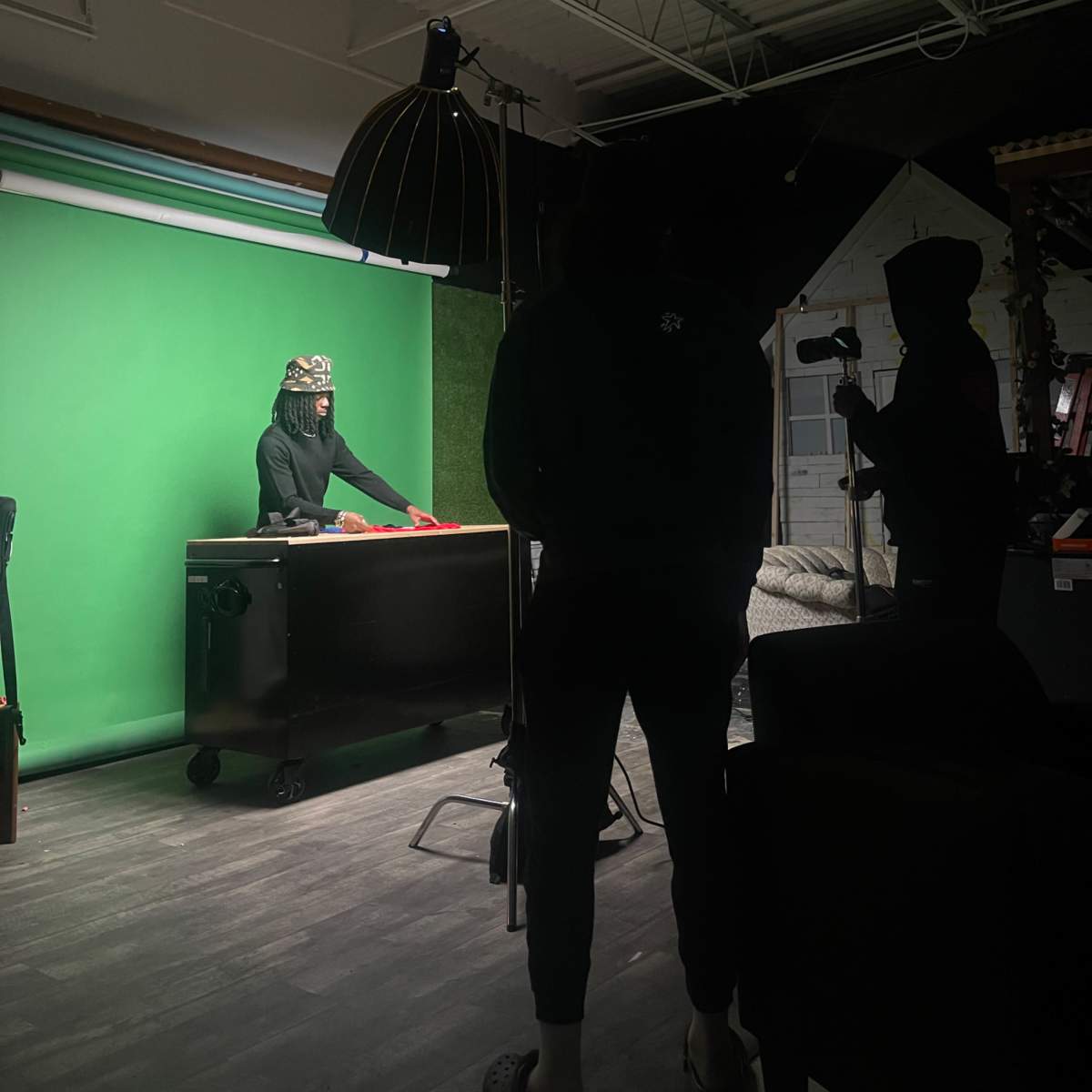 A person in a crown stands at a black table in front of a green screen, while two figures observe from the shadows.