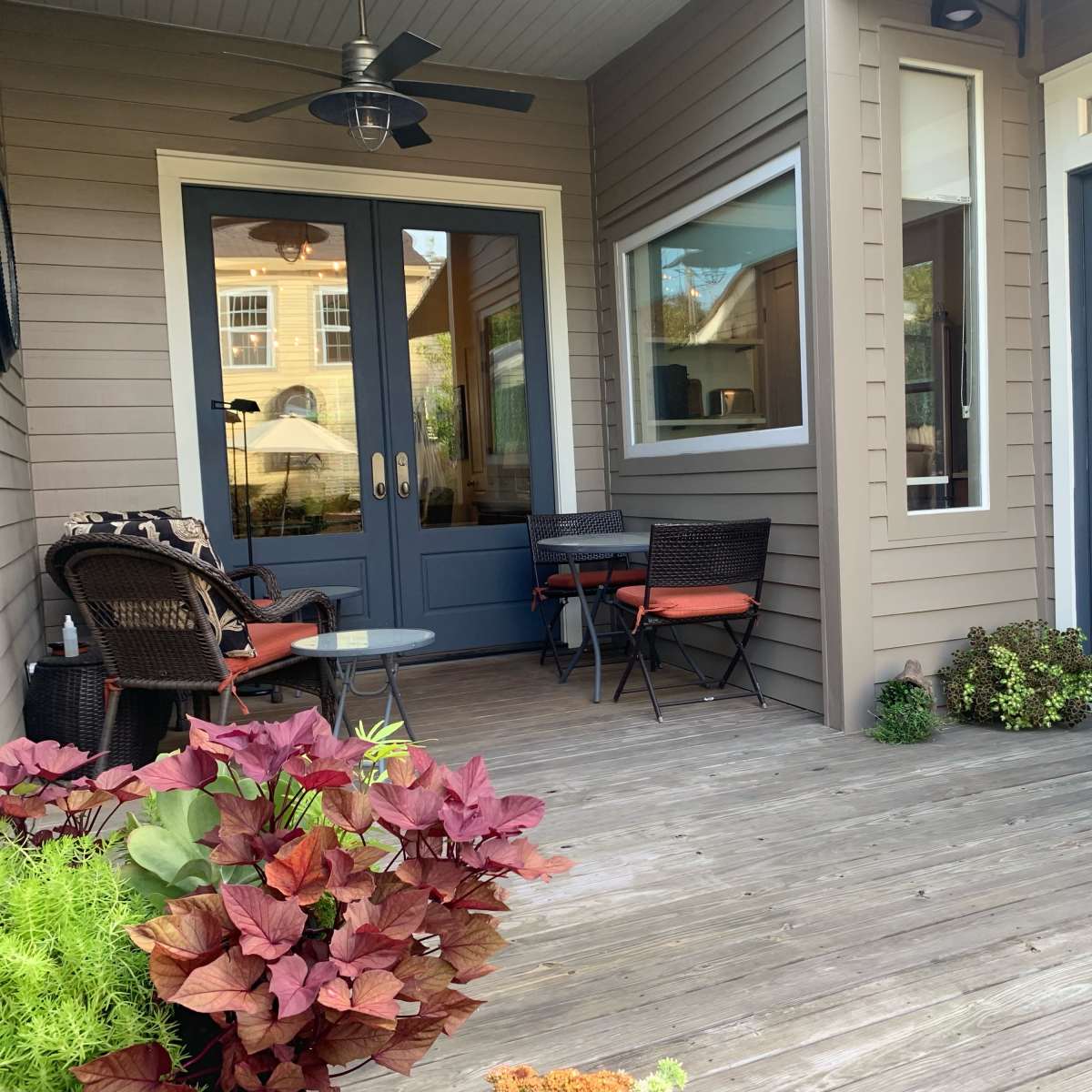 A wooden deck features two chairs and a small table beside a potted plant, with two blue doors leading into a building.