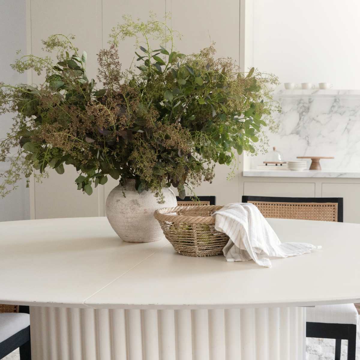 A large white dining table with a vase of greenery and a woven basket is set against a marble backdrop in a modern kitchen.