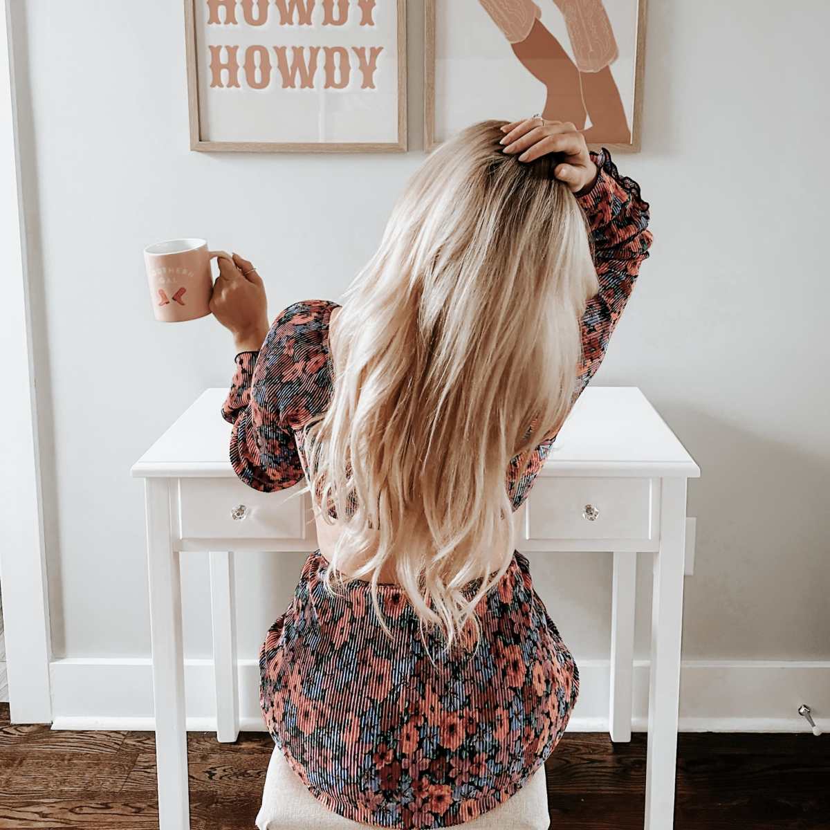A woman sits at a small white desk, holding a mug, with her back to the camera, and two framed art pieces on the wall that say "HOWDY" in bold text.