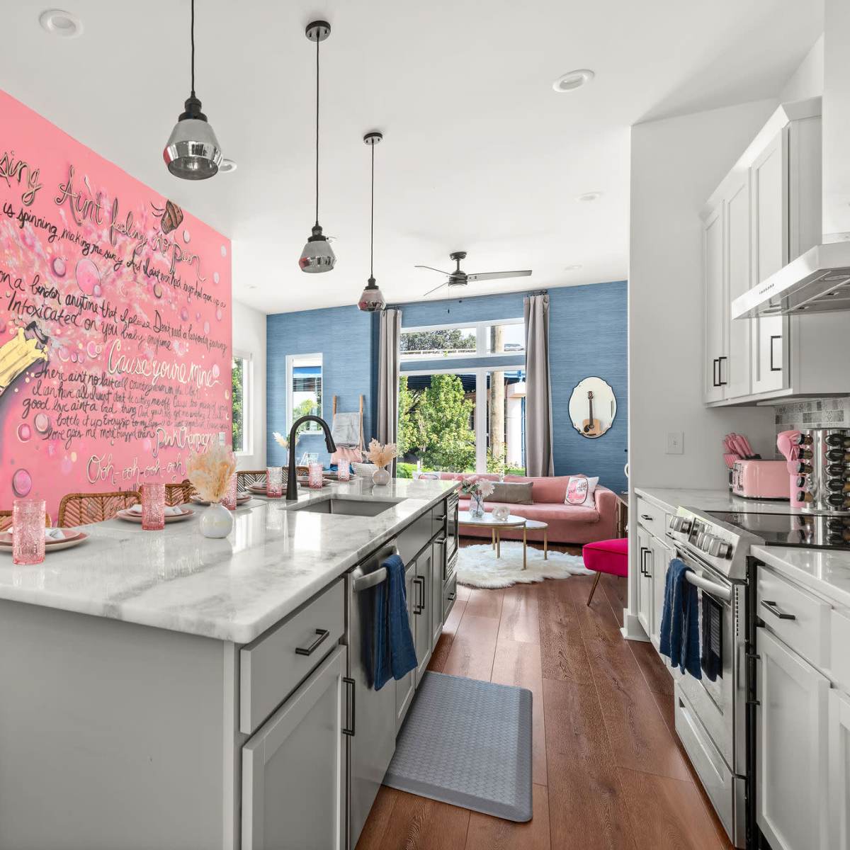 A modern kitchen features a pink mural of a wine bottle, with a marble countertop and a view into a brightly furnished living area.