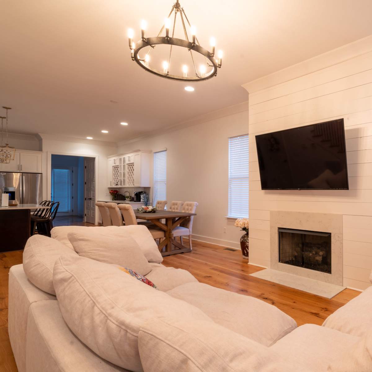 The living room features a light-colored sectional sofa facing a mounted television above a modern fireplace, with a dining area visible in the background.